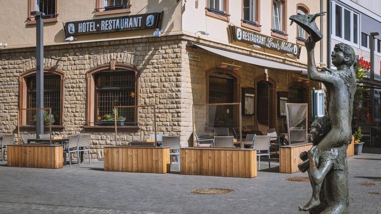 Ein Hotel-Restaurant mit einer gemütlichen Terrasse und einer Statue im Vordergrund. Die Fassade ist aus Stein und die Atmosphäre wirkt einladend.