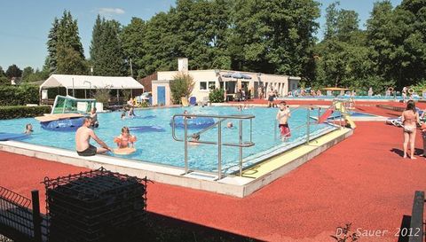 Une piscine avec plusieurs zones, où les gens nagent et se détendent. Il y a des chaises longues et un revêtement de sol rouge, entouré d'arbres.