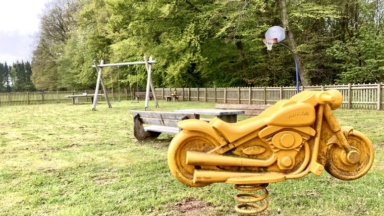 Ein Spielplatz mit einem Federspielzeug in Form eines Motorrads. Im Hintergrund sind Schaukeln und Bäume zu sehen.