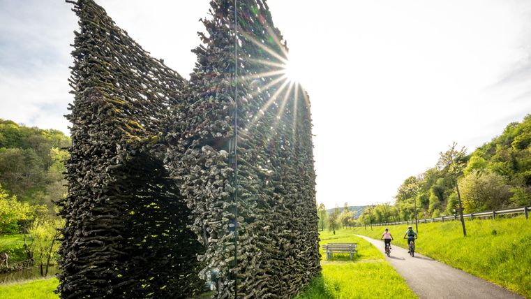 An artistic structure made of branches stands by the roadside. Two cyclists pass by as the sun rises.