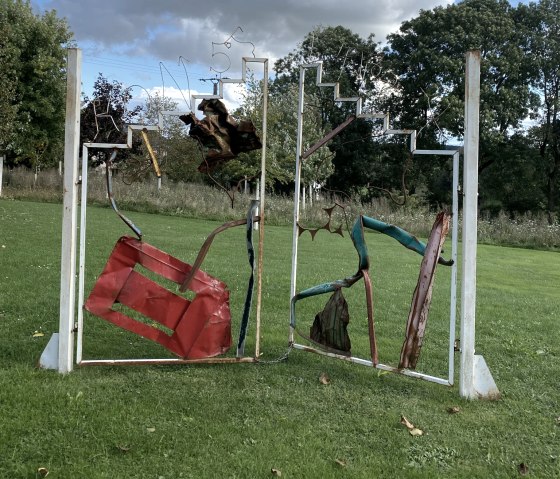 Metal sculpture on a green meadow, consisting of abstract forms and red elements, in front of a cloudy sky., © Felsenland Südeifel, Anna Carina Krebs