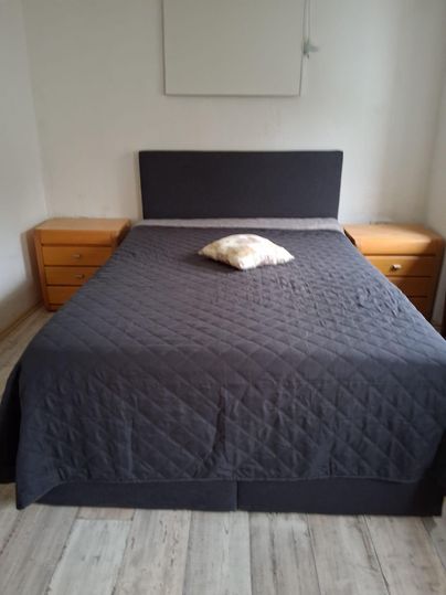 Une chambre simple avec un grand lit et deux tables de nuit en bois. Sur le lit, il y a un coussin au milieu.