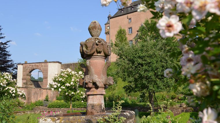Ein schöner Garten mit einer Statue im Vordergrund. Im Hintergrund ist ein historisches Gebäude und blühende Rosen zu sehen.
