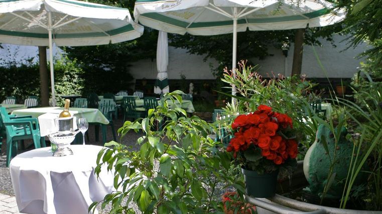 Un espace extérieur accueillant avec des plantes vertes et des fleurs rouges. Plusieurs tables et parasols créent une atmosphère conviviale.