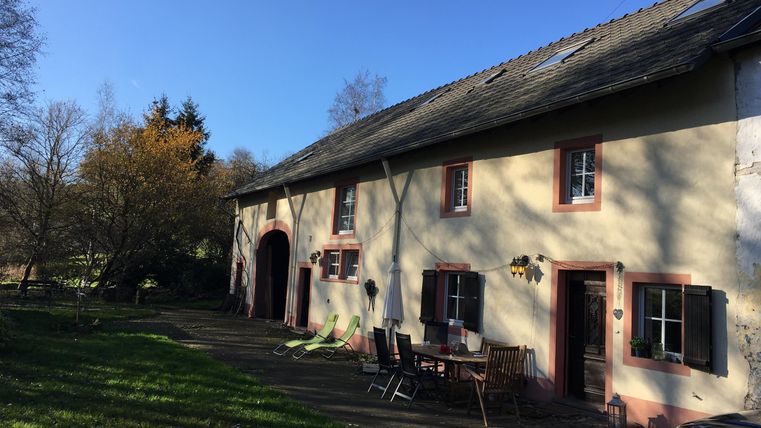 Ein rustikales Haus mit einem großen Garten. Der Himmel ist blau und es gibt Schatten von Bäumen.