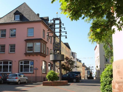 Eine ruhige Straße mit historischen Gebäuden und einem Glockenspiel in der Mitte. Die Sonne scheint und es gibt einige Autos geparkt.