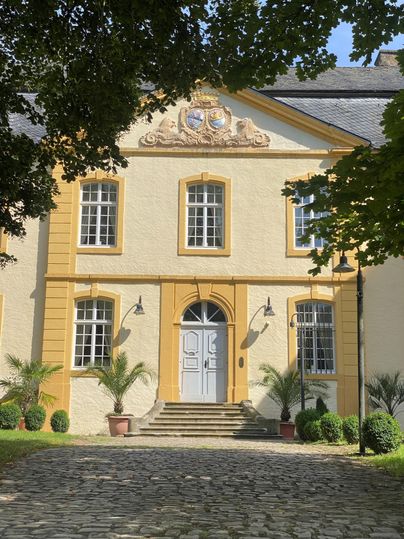 Ein elegantes Gebäude mit gelber Fassade und weißen Fenstern. Der Eingangsbereich ist von Pflanzen und einem gepflasterten Weg umgeben.