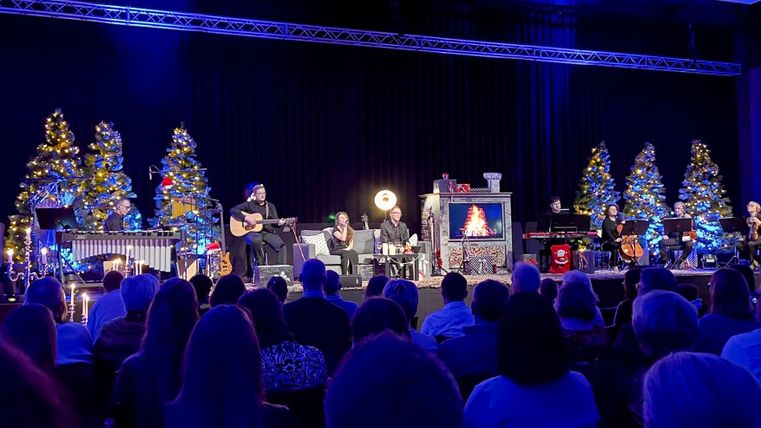 Eine festliche Bühnenaufführung mit Musikern und weihnachtlich geschmückten Bäumen. Das Publikum sitzt gespannt im Vordergrund.