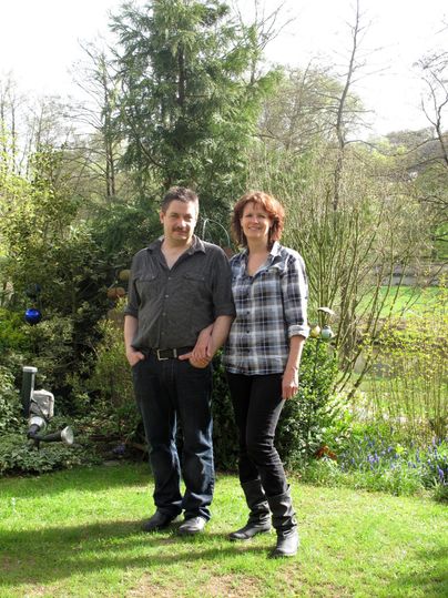Un couple se tient souriant dans le jardin. Autour d'eux, il y a des arbres et de l'herbe fraîche et verte.