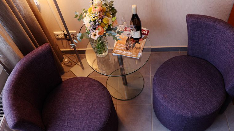 Deux fauteuils violets sont autour d'une table en verre avec un vase rempli de fleurs. Sur la table se trouvent un magazine et une bouteille de vin.