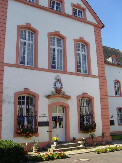 Ein historisches Gebäude mit einer weißen Fassade und rosa Details. Große Fenster und Blumen vor dem Eingang verleihen ihm einen einladenden Charakter.
