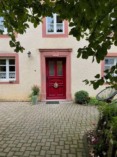 Een uitnodigende deur in het rood met een mooie deurkrans. Omringd door een geplaveide oprit en groene planten.