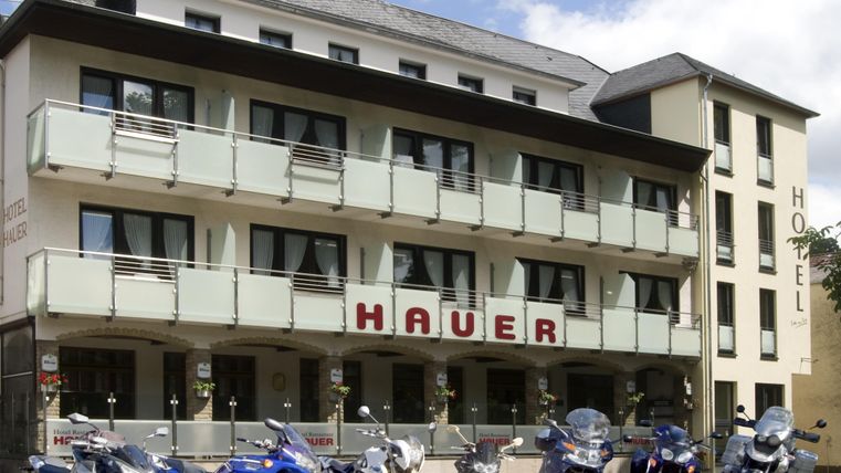 Un hôtel moderne avec l'inscription "HAUER" et plusieurs motos devant l'entrée. La façade est lumineuse et accueillante, entourée d'un ciel dégagé.