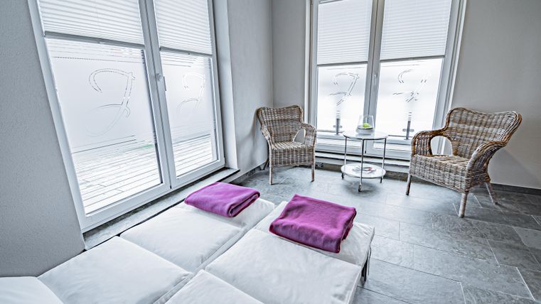 Ein ruhiger Raum mit zwei Liegen und lila Handtüchern. Zwei Korbstühle und ein kleiner Tisch stehen neben großen Fenstern.