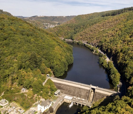 Luftaufnahme des Ourtalstausees mit bewaldeten Hügeln und einer Staumauer. Im Vordergrund sind Gebäude und Wege zu sehen., © Eifel Tourismus GmbH, D. Ketz