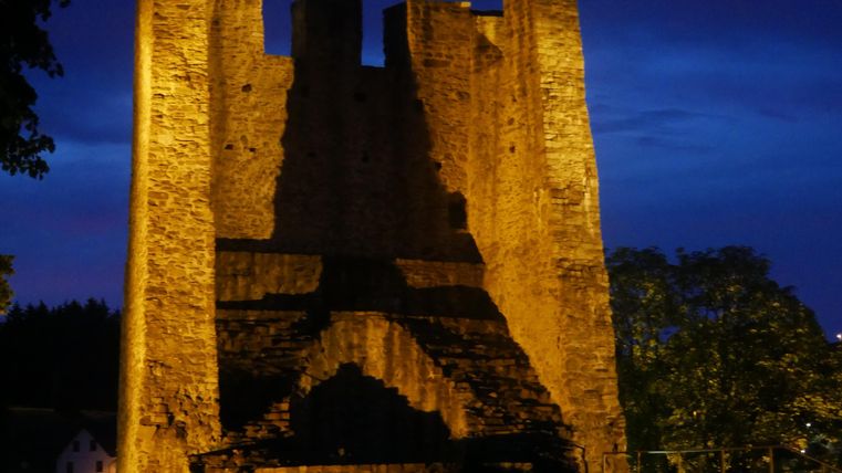 Eine alte Burg bei Nacht, beleuchtet von warmem Licht. Der Himmel ist dunkelblau und die Silhouette der Burg ist deutlich zu erkennen.