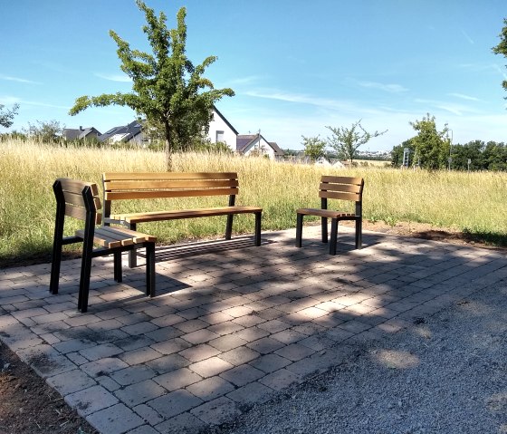 Three wooden benches stand on a paved square in a rural setting. Trees, a meadow and houses can be seen in the background., &copy; TI Bitburger Land