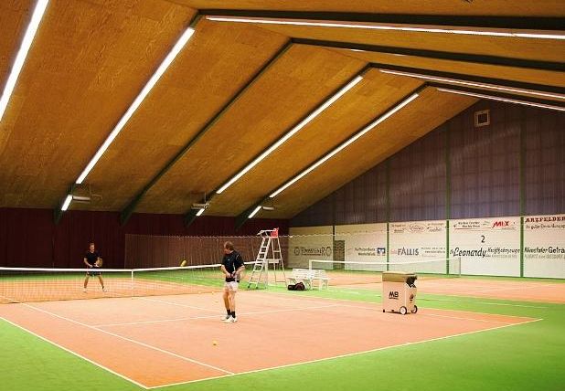 Eine Sporthalle mit einem Tennisplatz, auf dem zwei Spieler trainieren. Der Boden ist grün, und die Decke ist hoch und beleuchtet.