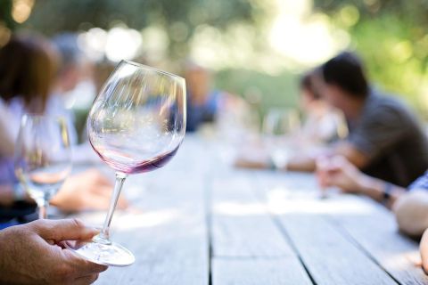 Ein Weinglas mit rotem Wein wird in der Hand gehalten. Im Hintergrund sind verschwommene Personen an einem langen Tisch zu sehen.