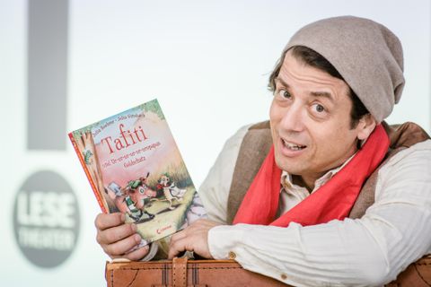 Ein Mann hält ein Buch mit dem Titel "Tafiti" in der Hand und lächelt freundlich in die Kamera. Er trägt einen grau-weißen Hut und einen roten Schal, während er vor einem neutralen Hintergrund steht.