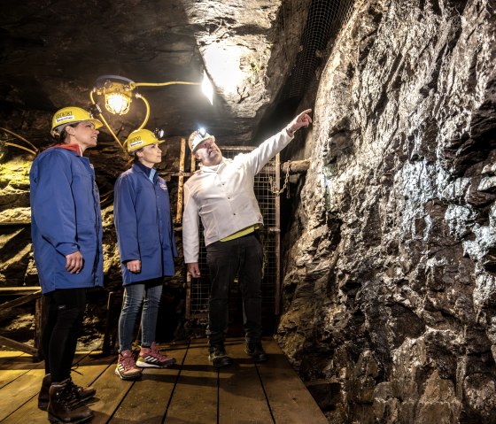 Drei Personen in Schutzkleidung stehen in einem Bergwerk. Eine Person zeigt auf die Wand, die von einer Lampe beleuchtet wird., &copy; Eifel Tourismus GmbH, Dominik Ketz