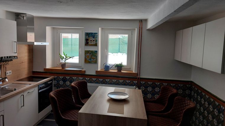 Een moderne keuken met een houten tafel en stoelen. Twee ramen laten veel licht binnenkomen en er zijn enkele planten en afbeeldingen aan de muren.