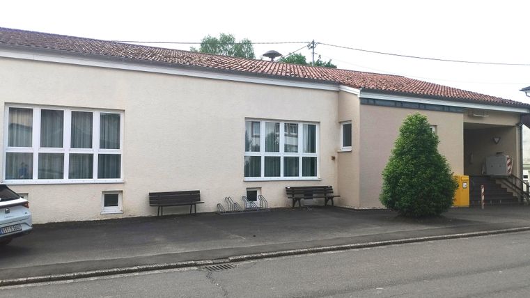 Ein einfaches Gebäude mit weißer Fassade und mehreren Fenstern. Draußen stehen Bänke und ein kleiner Baum ist sichtbar.