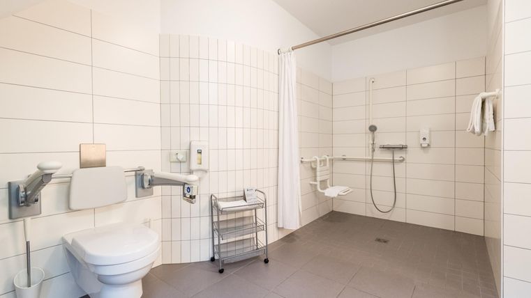 Een rolstoeltoegankelijke badkamer met een toiletruimte en een douche. De muren zijn licht betegeld en er zijn leuningen voor extra veiligheid.