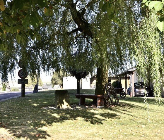 Ein schattiger Rastplatz mit Bank unter einem Baum neben einer Straße in Stockem, Naturpark Südeifel. Ein Auto steht in der Einfahrt eines Hauses., © TI Bitburger Land