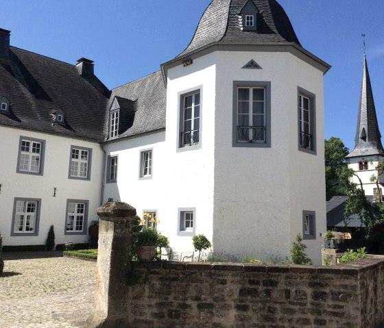 White castle with a gray roof and tower, surrounded by a garden and stone wall, under a blue sky., © Fellmann / TI Bitburger Land