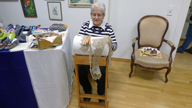 Eine ältere Dame sitzt an einem Tisch und arbeitet an einer Klöppelarbeit. Neben ihr befindet sich ein Stuhl und einige Materialien auf dem Tisch.