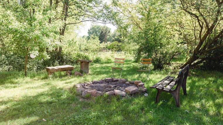 Ein gemütlicher Garten mit einer Feuerstelle und mehreren Sitzgelegenheiten. Umgeben von grünen Bäumen und viel Licht.