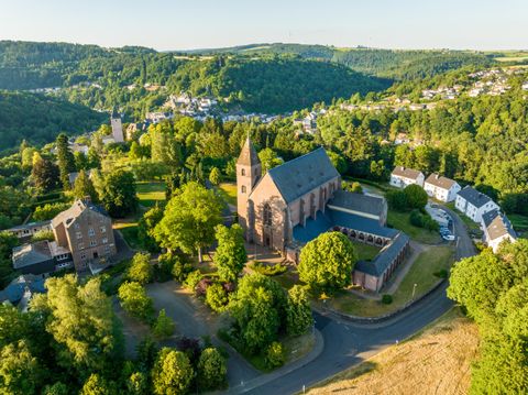 Ein malerisches Dorf umgeben von grünen Hügeln und Bäumen. Im Vordergrund ist eine historische Kirche sichtbar, die von Natur umgeben ist.