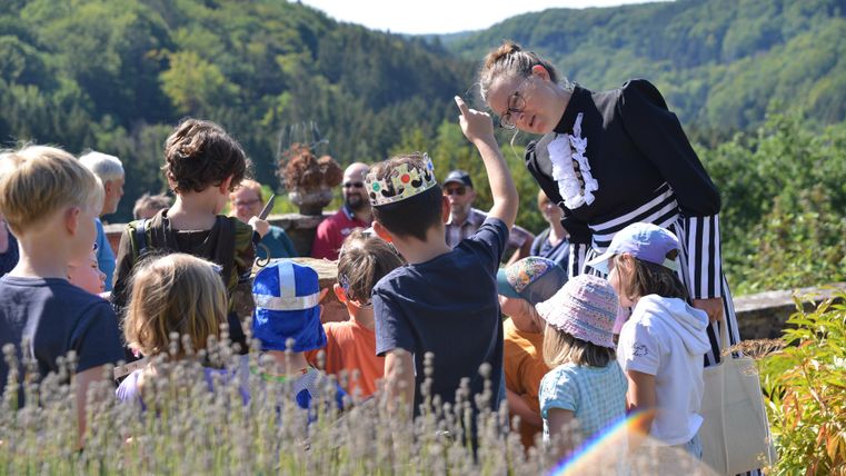 Eine Gruppe von Kindern hört einer Frau in Kostüm aufmerksam zu. Im Hintergrund sind grüne Wälder und eine sonnige Landschaft zu sehen.