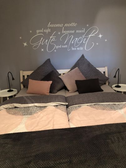 Ein gemütliches Schlafzimmer mit einem großen Bett und vielen Kissen. An der Wand steht der Schriftzug „Gute Nacht“.