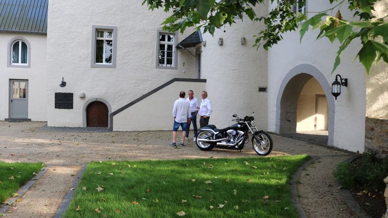 Zwei Männer unterhalten sich vor einem eleganten Gebäude. Ein Motorrad steht neben ihnen auf der gepflasterten Fläche.