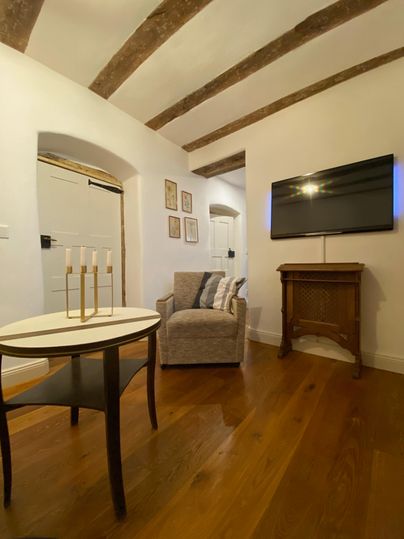 Een gezellig woonkamer met een fauteuil, een tafeltje en een televisie aan de muur. De houten vloeren en plafondbalken geven de ruimte een warme sfeer.