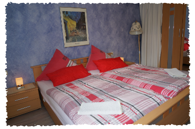 Une chambre confortable avec un lit double, des coussins rouges et une couette colorée. Sur le mur, il y a un tableau, et il y a une lampe de chevet ainsi qu'une armoire.