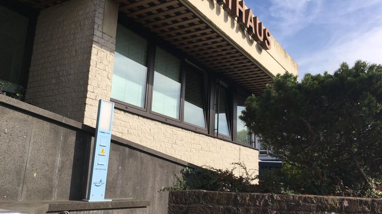 A modern town hall building with large windows and an inscription. In the foreground, bushes and a staircase can be seen.