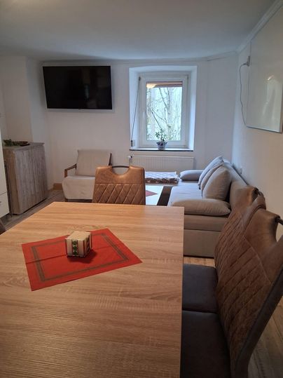 Un espace de vie confortable avec une table en bois et une télévision au mur. La pièce est lumineuse et possède de grandes fenêtres donnant sur l'extérieur.
