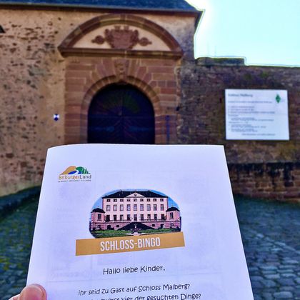 Ein Handzettel für "Schloss-Bingo" wird vor einem historischen Schloss gehalten. Im Hintergrund ist die Eingangstür des Schlosses und ein Informationsschild sichtbar.