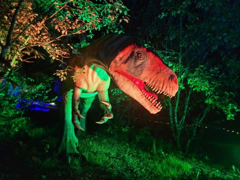 Ein lebensechter Dinosaurier, der zwischen Bäumen steht, mit grünem und rotem Licht beleuchtet. Die Szene vermittelt eine mystische und spannende Atmosphäre.