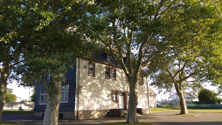 Ein zweistöckiges Haus umgeben von großen Bäumen. Die sonnige Atmosphäre vermittelt eine ruhige und einladende Stimmung.