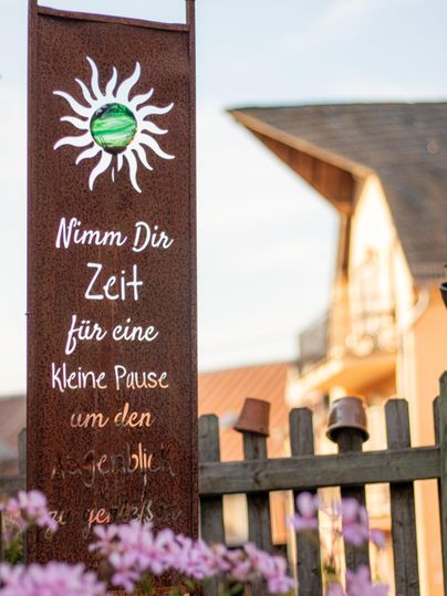 Ein dekoratives Schild mit der Aufschrift „Nimm Dir Zeit für eine kleine Pause“ steht vor einem kaufspeziellen Hintergrund. Bunte Blumen sind im Vordergrund sichtbar.
