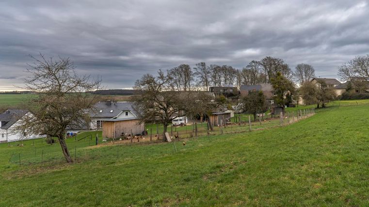 Eine malerische ländliche Szene mit Häusern und einem grünen Feld. Der Himmel ist bewölkt, und Bäume umgeben die Gebäude.