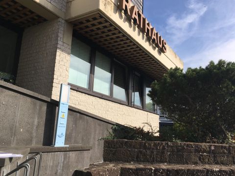 A modern town hall building with large windows and an inscription. In the foreground, bushes and a staircase can be seen.