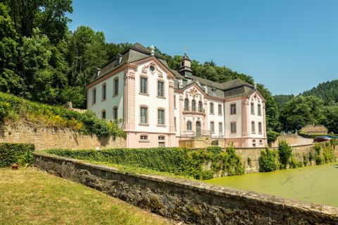 Ein imposantes historisches Gebäude mit weißen Wänden und roten Akzenten. Es ist von üppigem Grün und einem ruhigen Teich umgeben.