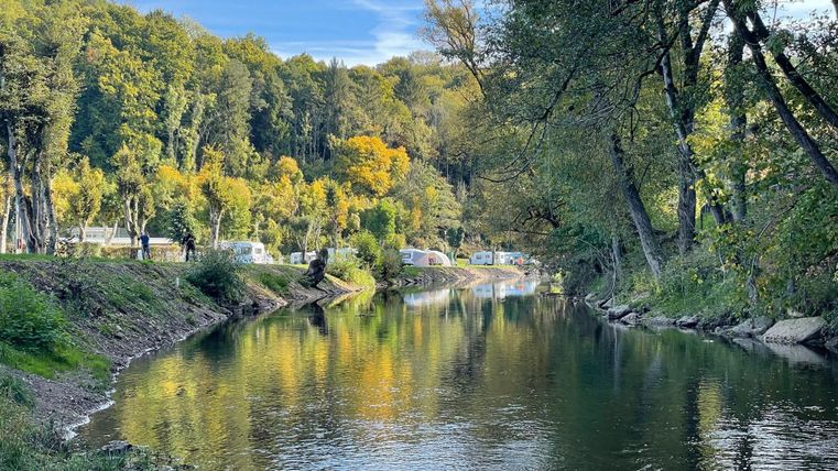 Ein ruhiger Fluss umgeben von bunten Bäumen im Herbst. Im Hintergrund sind einige Wohnwagen entlang des Ufers zu sehen.