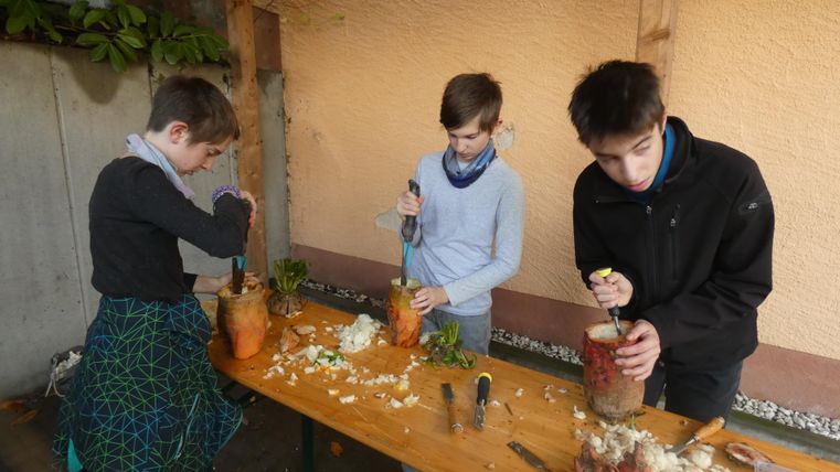 Drei Jungen arbeiten an einem Tisch, um Gemüse zu schneiden und zuzubereiten. Der Tisch ist mit verschiedenen Zutaten und Werkzeugen bedeckt.