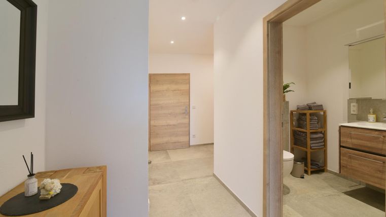 Een heldere en moderne gang met een houten kast en een spiegel. Rechts ziet men een toegang tot een badkamer.